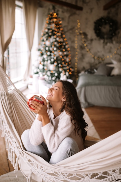 Smiling dark-haired girl dressed in beige sweater and pants holds a red cup sitting in a hammock in a cozy decorated room with a New Year tree . .