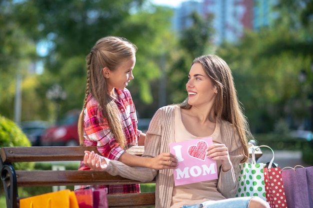 Smiling daughter presenting card for her mom sitting on park bench