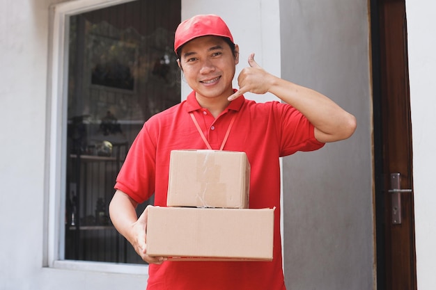 Smiling delivery man making a call me gesture while carrying stack of packages outdoor