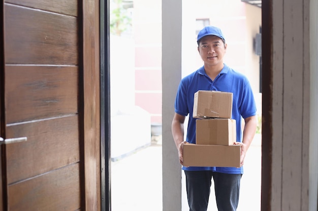 Smiling deliveryman in blue uniform holding pile of boxes in front of the door.