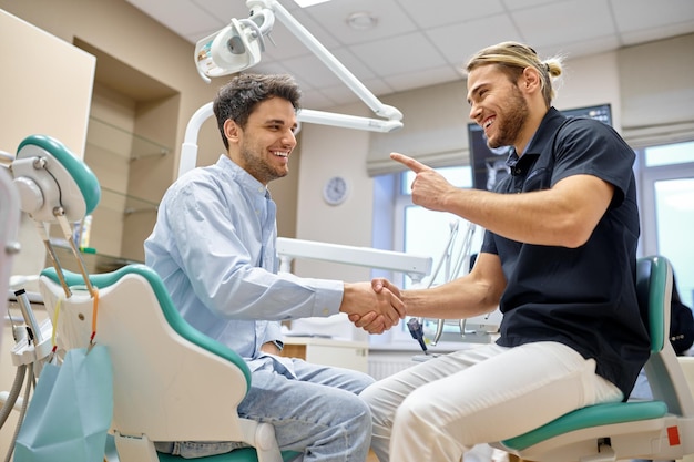 Smiling dentist shaking hand with male patient. Stomatologist giving teeth treatment and care advice