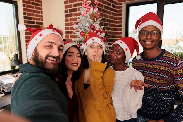 Smiling diverse men and women colleagues wearing santa hats posing for selfie together in office with christmas decorations Cheerful coworkers taking group photo at new year corporate party