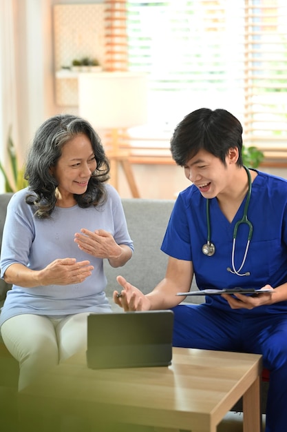 Smiling doctor in blue medical uniform showing good health test result on digital tablet to happy senior female patient during home visit