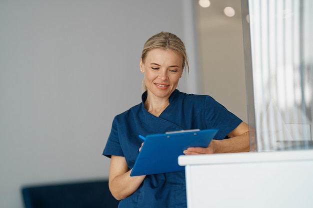 Smiling doctor making notes in clipboard standing near reception in clinic hall