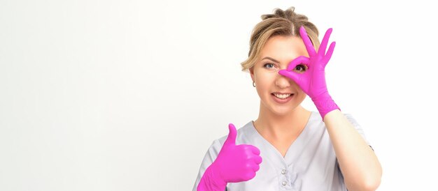 Smiling doctor oculist caucasian woman wearing pink rubber gloves in uniform showing ok sign covering the eye and thumb up gesture against a white background.