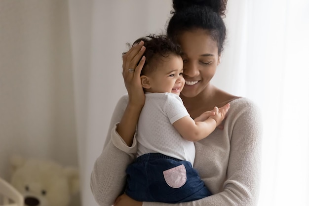 Smiling dreamy African American young mother holding cute little daughter close up looking to aside at window dreaming about good future planning caring happy mum hugging toddler girl