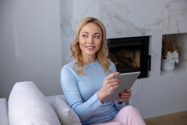 Smiling dreamy beautiful Caucasian blonde woman sitting on the cozy sofa with her tablet computer