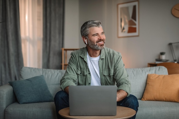 Smiling elderly european man with beard in wireless headphones typing on laptop and looking at empty