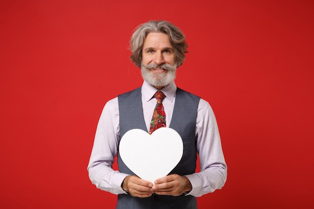 Smiling elderly gray-haired mustache bearded man in classic shirt vest colorful tie posing isolated on red background. People lifestyle concept. Mock up copy space. Holding white blank empty heart.