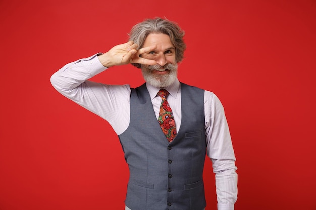 Smiling elderly gray-haired mustache bearded man in classic shirt vest and colorful tie posing isolated on red background in studio. People lifestyle concept. Mock up copy space. Showing victory sign.