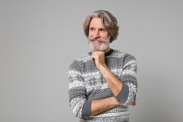 Smiling elderly gray-haired mustache bearded man in knitted sweater posing isolated on grey wall background, studio portrait. People lifestyle concept. Mock up copy space. Put hand prop up on chin.