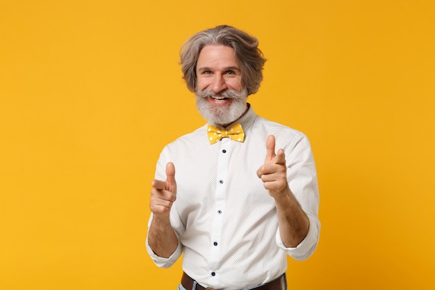 Smiling elderly gray-haired mustache bearded man in white shirt bow tie posing isolated on yellow background in studio. People lifestyle concept. Mock up copy space. Pointing index fingers on camera.