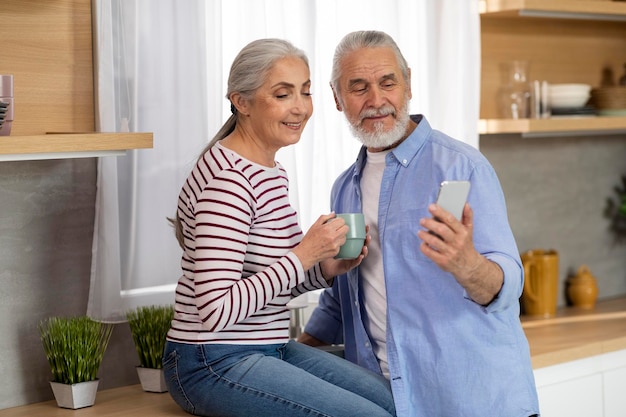 Smiling Elderly Husband And Wife Relaxing In Kitchen With Smartphone And Coffee