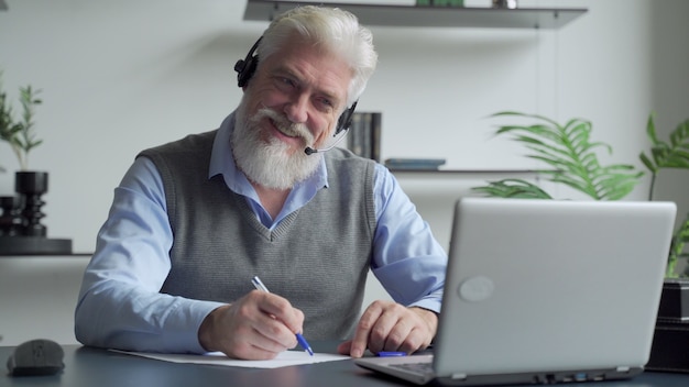 Smiling elderly man with a gray beard wearing headphones having a video call on a laptop happy elderly man