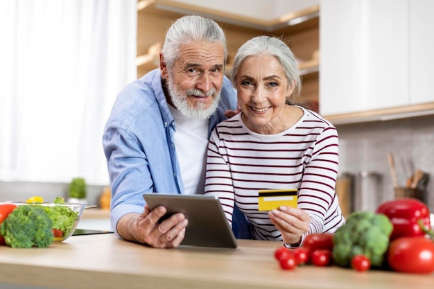 Smiling Elderly Spouses With Digital Tablet And Credit Card In Kitchen