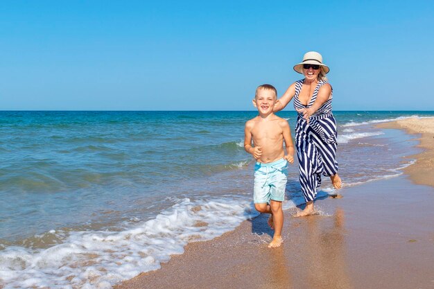 Smiling elderly woman in a dress and hat with a boy run along the sandy beach near the sea on a sunny day Love tenderness and an active lifestyle