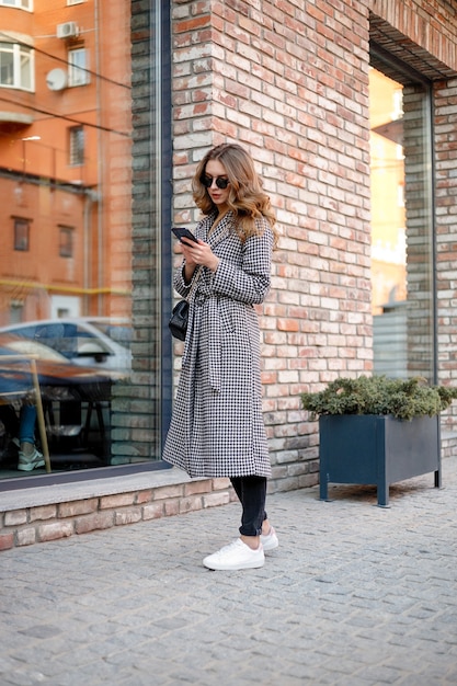 Smiling elegant woman in trench coat walking and drinking coffee from paper cup . smiling woman with curly hair look at camera smile walking slow motion face sunset beautiful lady outdoor closeup cute