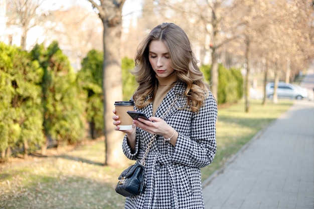 Smiling elegant woman in trench coat walking holding mobile and coffee