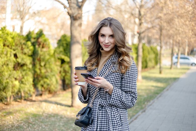 Smiling elegant woman in trench coat walking holding mobile and coffee