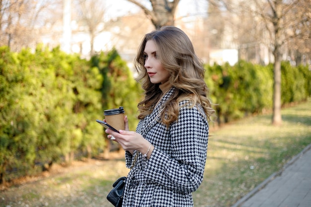 Smiling elegant woman in trench coat walking holding mobile and coffee