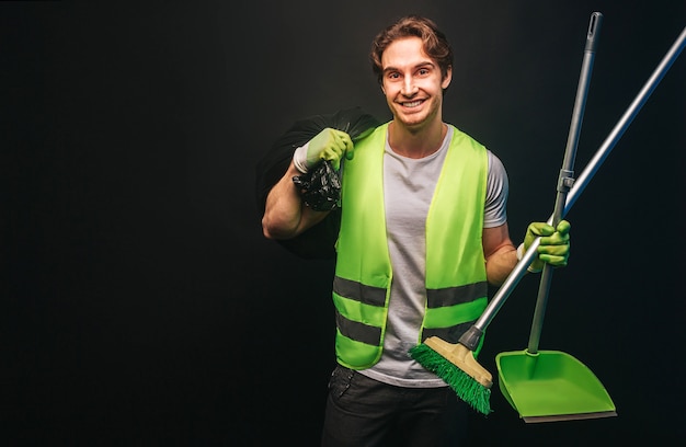 Smiling european cleaner hold rubbish bag, broom and scoop. Young handsome man wear green uniform and rubber gloves. Concept of cleaning service. Isolated on dark background. Studio shoot. Copy space