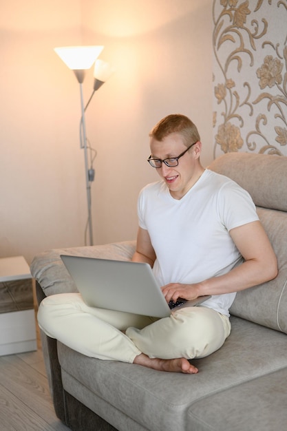 Smiling European man in glasses dressed in light clothing uses a laptop for online work sits on the sofa at home