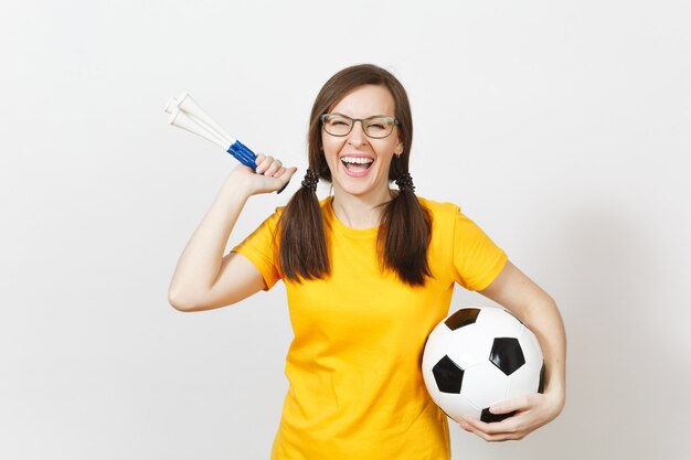 Smiling European woman, two fun pony tails, football fan or player in glasses, yellow uniform hold football pipe, ball isolated on white background. Sport, football, health, healthy lifestyle concept.