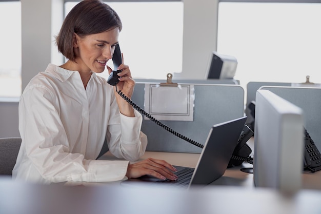 Smiling european woman worker talk phone with client sit on her workplace in office