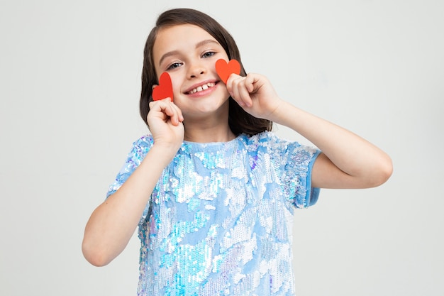 Smiling european young girl holding two cards in the shape of a heart for valentines day on a light