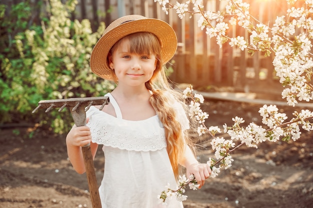 Smiling farmer girl in a hat in the garden is engaged in gardening, a rake for harvesting leaves in her hands. Spring flowering of apricots. The garden is in bloom. Sunlight on background