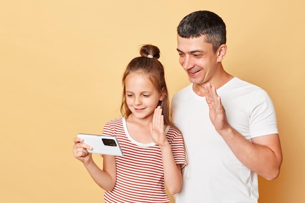 Smiling father and daughter wearing casual tshirts standing isolated over beige background having online video call waving hands saying hello or bye