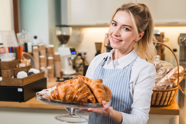 Smiling female baker holding baked croissant in the cake stand