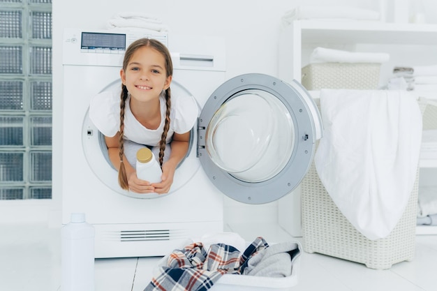 Smiling female child with glad expression poses in washing machine dressed in white t shirt holds detergent looks gladfully at camera surrounded with much clothes and linen does housework