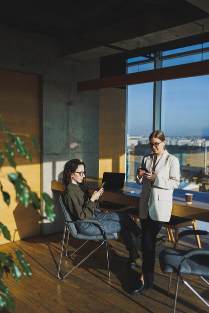 Smiling female colleagues collaborating indoors at table woman wearing glasses and using laptop and showing partner Business relations of serel colleagues in the office Team work of colleagues