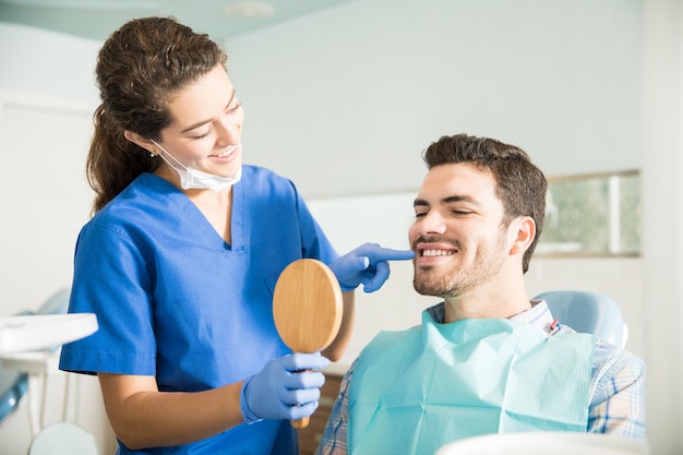 Smiling female dentist showing mirror to mid adult man after treatment at dental clinic