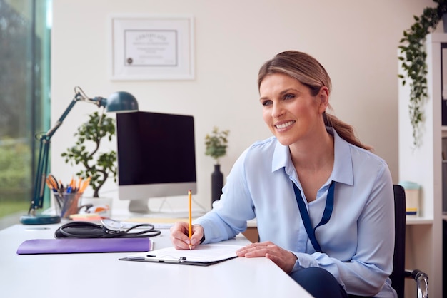 Smiling Female Doctor Or GP Sitting At Desk In Office Writing Notes On Clipboard