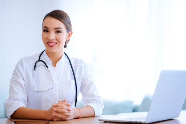 Smiling female doctor with stethoscope wearing laboratory coat works in hospital and using laptop