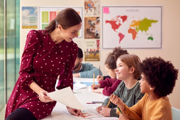 Smiling Female Elementary School Teacher Working At Desk In Classroom With Students