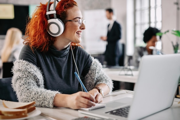 Smiling female entrepreneur having fun while working in the office and listening favorite music on headphones There are people in the background