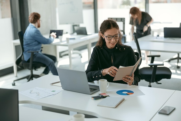 Photo smiling female entrepreneur holding note pad while working on laptop sitting in modern coworking