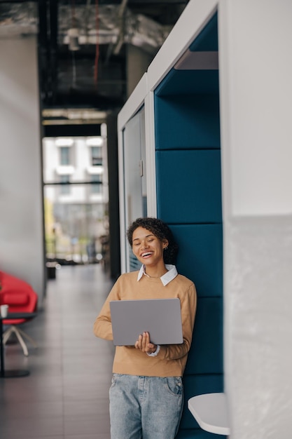 Photo smiling female entrepreneur using laptop while standing on office background and looks camera