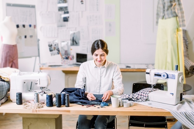 Smiling female fashion designer sitting at office desk. Young woman sewing with sewing machine at studio while sitting at her working place.