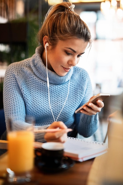 Smiling female freelancer writing do to list in her weekly planner while using mobile phone in cafeteria