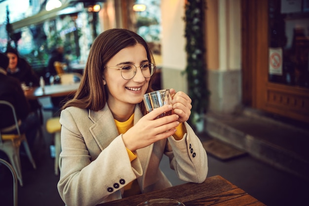 Smiling female influencer sitting on terrace of bar, holding cup of tea and warming up hands.