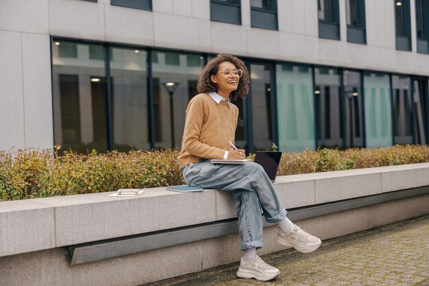 Photo smiling female manager working on laptop and making notes sitting on background of office building