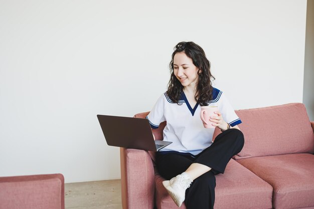 Smiling female student sitting on sofa in bright cafe with coffee looking at laptop Remote work with a laptop in a cozy work cafe