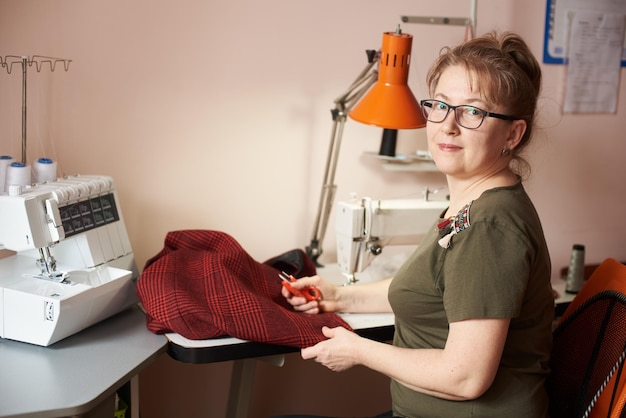 Smiling female tailor on sewing process of skirt holding scissors Overlock and sewingmachine on background Side view