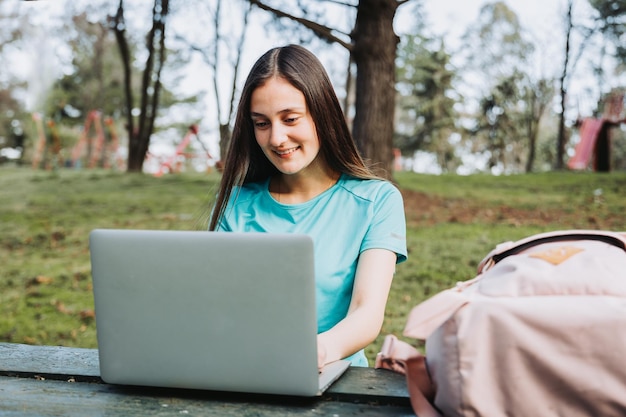 Smiling female university student using her laptop in campus natural park. E-learning concept.