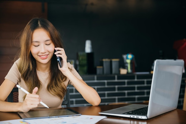 Smiling female working laptop computer and take note on digital tablet at home office, Happy freelance Asian woman sitting at cafe call mobile phone talking and asking about something, financial