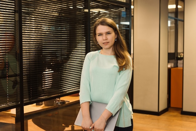 Smiling freelancer woman working at co-working or creative space, standing with laptop on the corridor of office